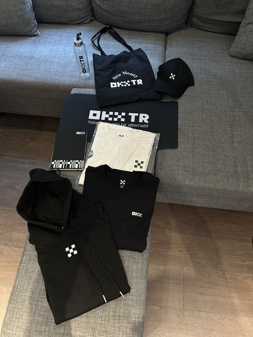 A collection of OKX TR branded merchandise, including a black t-shirt, hoodie, cap, tote bag, water bottle, notebook, and packaged shorts, is arranged on a gray couch and floor mat. The text "OKX TR" and "Take Money" are visible on several items, with the post thanking the OKX TR family for the gifts and announcing a giveaway for followers.