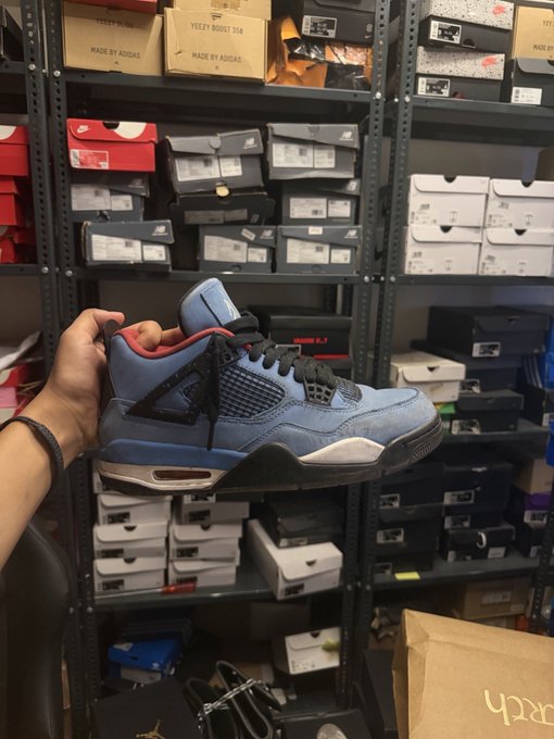 A hand holds up a blue and red Air Jordan sneaker in front of shelves stocked with various shoe boxes in what appears to be a sneaker store.