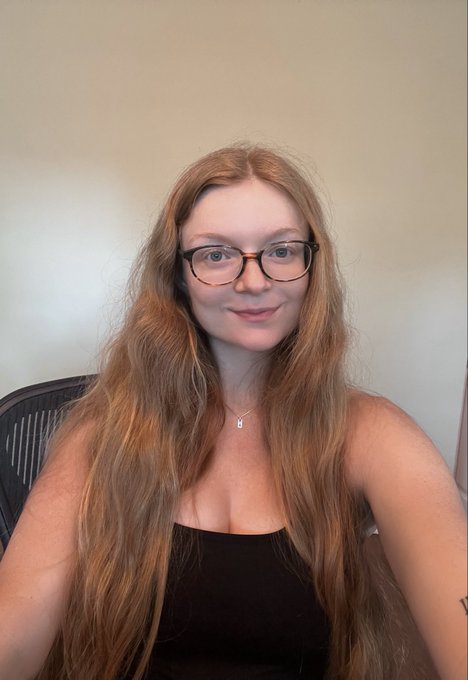 A young woman with long reddish-brown hair and tortoiseshell glasses, wearing a black strapless top and a small necklace, sitting in front of a light background, possibly in an office setting.