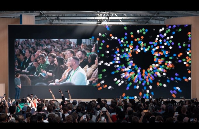 A presenter on stage stands before a large audience and a giant screen displaying both the crowd and colorful dot graphics.