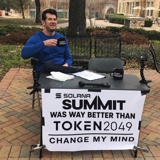 A man sits at an outdoor table holding a mug that reads "SUPER CHOWDER," with a sign in front of him stating "SOLANA SUMMIT WAS WAY BETTER THAN TOKEN2049 CHANGE MY MIND."