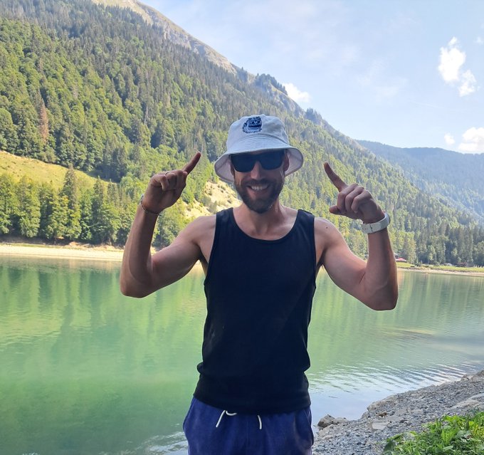 A smiling man wearing a gray bucket hat, sunglasses, and a black tank top points both index fingers upward while standing in front of a green lake surrounded by forested mountains.