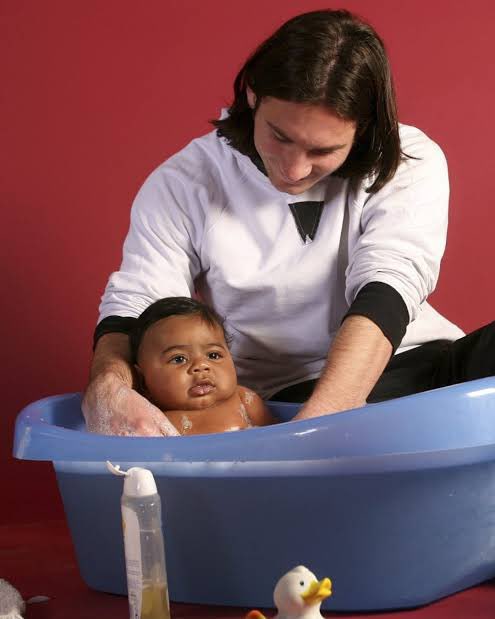 A man in a white long-sleeve shirt bathes a baby in a blue plastic tub against a red background, with baby products and a rubber duck visible in the foreground.