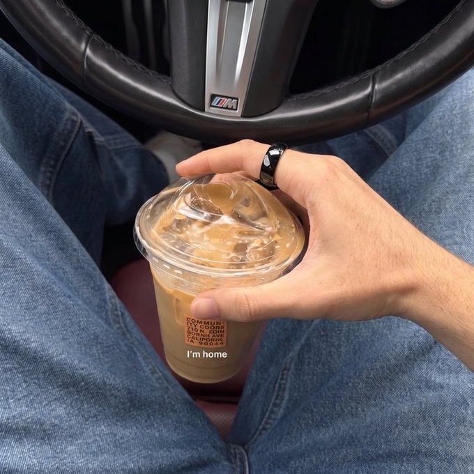 A first-person view from the driver's seat shows a hand holding an iced coffee in a clear plastic cup labeled "I'm home" from Community Goods Cold Brew.