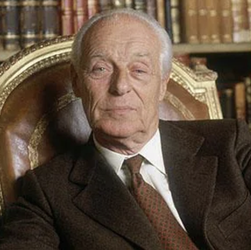 A portrait photograph of an elderly man in a dark suit and patterned tie, seated in an ornate chair with bookshelves visible in the background.