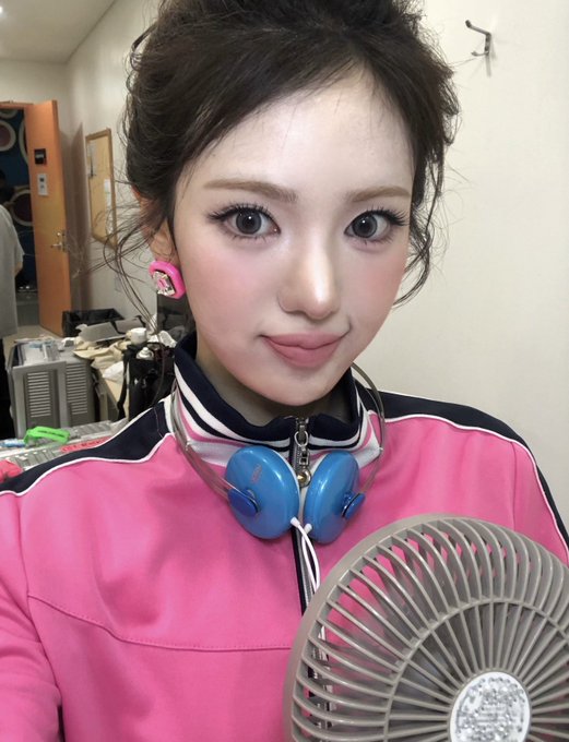 A young woman wearing a pink tracksuit jacket poses for a selfie in front of a small fan, with blue headphones around her neck.