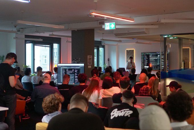 A presenter stands before a packed audience in a modern event space, with two large screens displaying code and data visualizations flanking the stage. The "Veni, vidi, vici" caption ("I came, I saw, I conquered") frames the scene as a triumphant tech talk or developer meetup, capturing the moment of the speaker "conquering" the attentive crowd.