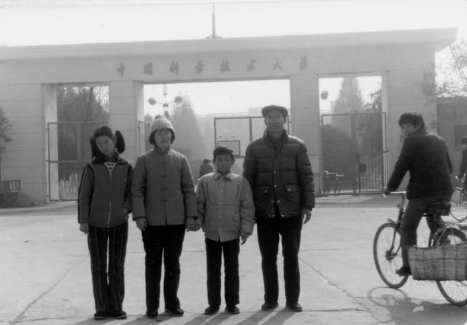A black-and-white photograph shows a family of four posing in front of the gate of the University of Science and Technology of China (中国科学技术大学), with Chinese characters visible across the top of the entrance archway and a man riding a bicycle passing by on the right. The image likely captures CZ's father, an older man in a winter jacket, with his family during his early academic days at the university following the restoration of China's college entrance examination system in 1978.