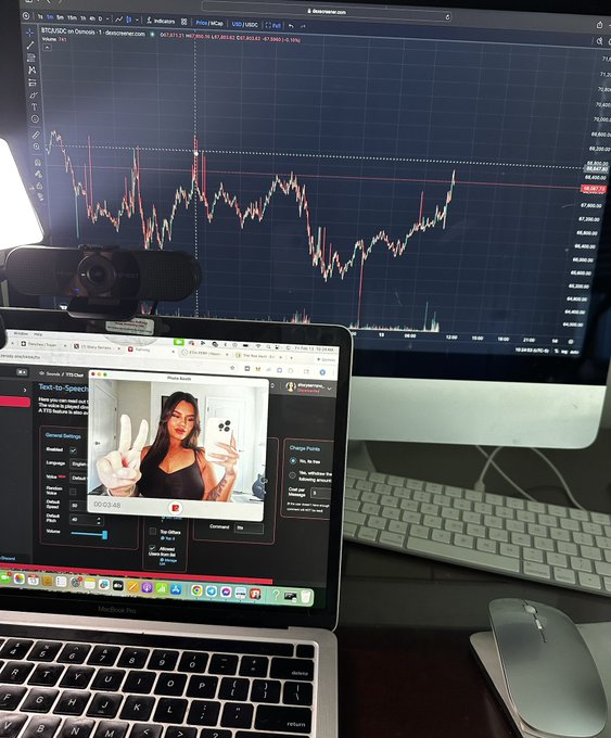 A desk setup showing a large monitor displaying a BTC/USD candlestick chart on TradingView alongside a laptop running a StreamYard broadcast featuring a woman holding up two fingers.