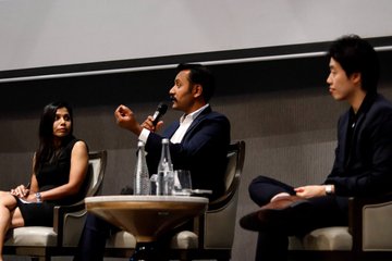 Three people sit on a panel discussion stage, with the central man in a suit speaking into a microphone while holding a pen, flanked by two other individuals.