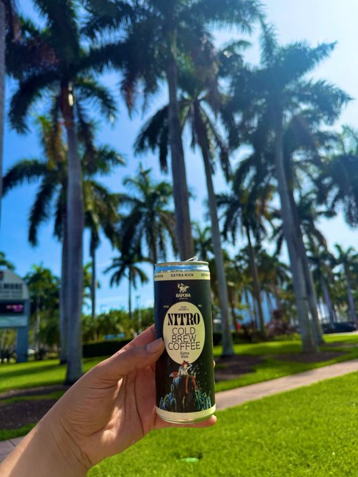The image features a person holding up an aluminum can of coffee, which has "Xtiro" written on it. The scene takes place outdoors with palm trees visible in the background. There are also other people present in the image, but they are not the main focus. The photo is related to the post because it shows a person holding a can of Xtiro Coffee, which could be an NFT project or a product from the blockchain.