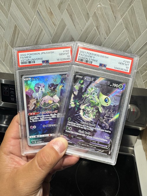 A hand holds two PSA-graded Pokemon cards in plastic slabs: a 2022 Pokemon Jpn SWSH Faimew VSTAR Universe (#183, Gem Mint 10) and a 2021 Pokemon SWSH Celebi V Fusion Strike (#245, Gem Mint 10).