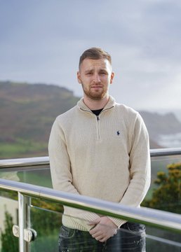 A young man wearing a cream-colored half-zip sweater stands with hands clasped in front of a railing with a scenic mountainous landscape in the background.