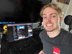 A young man sits in front of a dual-monitor computer setup displaying chat windows, wearing a dark gray t-shirt, with a small yellow duck plushie visible on the left monitor.