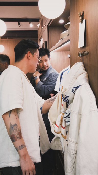 Two men with visible tattoos browse hanging garments in a warmly lit, wood-paneled boutique, with one examining a cream-colored jacket while a clerk assists. The casual, fashion-forward setting aligns with the minimalist "gM" caption, suggesting a stylish shopping moment.