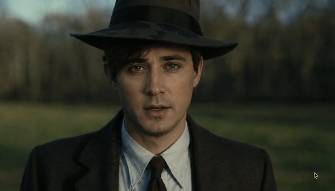 A close-up portrait of a man wearing a dark fedora, suit, white shirt, and dark tie, set against a blurred background of trees.