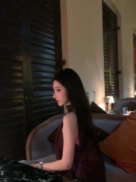 A side profile of a young woman with long dark hair wearing a dark sleeveless dress, sitting on a couch in an elegant indoor setting with louvered doors.