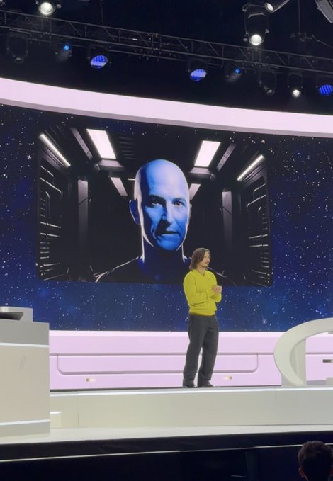A woman in a yellow top presents on stage at the Robinhood keynote, standing in front of a large screen displaying an image of Coinbase CEO Brian Armstrong—identified in the post as the real person behind the "Vrax" character.