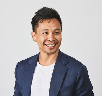 A professional portrait of a smiling man with short dark hair wearing a navy blue blazer over a white t-shirt against a light gray background.