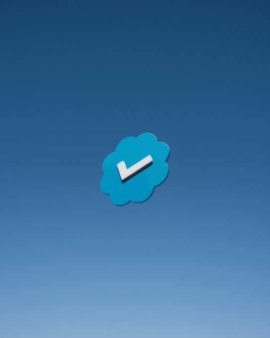 A 3D-rendered blue cloud-shaped icon with a white checkmark sits against a gradient blue sky background, resembling a Twitter/X verification badge.