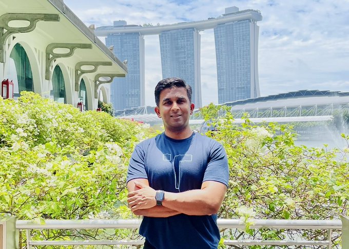 A man in a blue t-shirt with arms crossed stands in front of greenery with the Marina Bay Sands hotel visible in the background in Singapore.