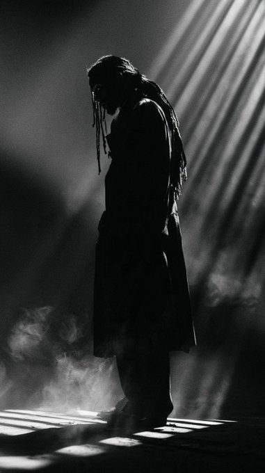 A black-and-white silhouette of a person with long hair standing in dramatic rays of light amid smoke on a stage.