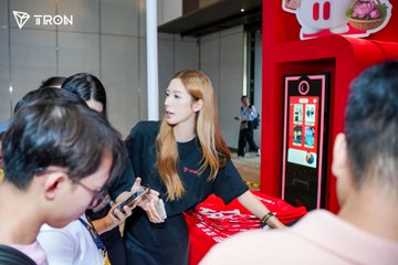 A young woman with long blonde hair stands at the TRON booth during the Conviction2026 event in Ho Chi Minh City, engaging with attendees. The "TRON" logo is visible in the upper left corner, while she interacts with visitors near a red gaming display.