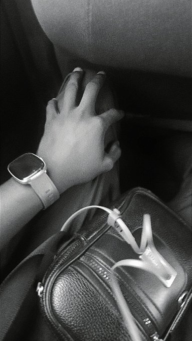 A black-and-white photo shows a hand with painted nails wearing a smartwatch resting near a bag with tangled wired earphones, likely taken during a mundane commute. The casual accessories and text suggest someone capturing an ordinary moment before or after sharing workplace news.