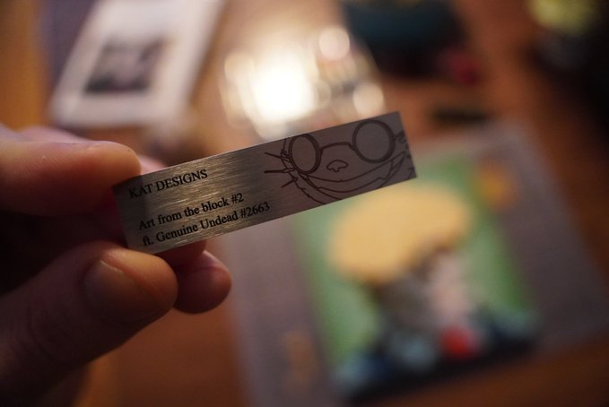 A close-up of a hand holding a small metal label that reads "KAT DESIGNS, Art from the Block #2, ft. Genuine Undead #2663" with an engraved character illustration, set against a blurred background with bokeh lights.