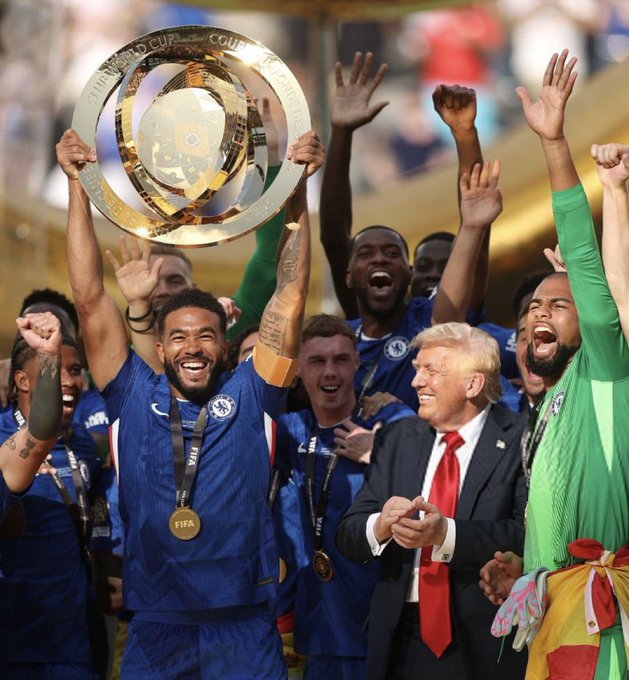 Chelsea players celebrate winning the FIFA Club World Cup trophy, with one player hoisting the gold trophy while former US President Donald Trump stands smiling nearby in a suit and red tie.