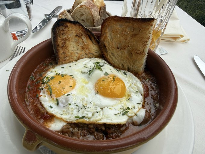 A clay dish containing two sunny-side-up eggs in tomato sauce, served with two slices of toasted bread on a white tablecloth.