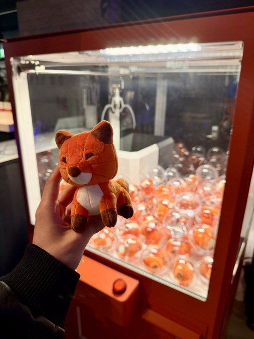 A hand holding an orange fox plush toy in front of a claw machine filled with clear capsule toys containing similar orange items.