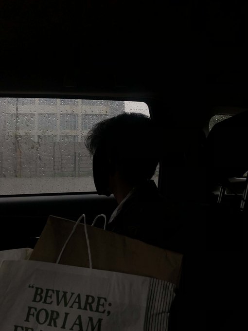 A silhouetted figure sits in the back of a vehicle next to a rain-streaked window, with a paper bag reading 'BEWARE: FOR I AM' visible in the foreground.