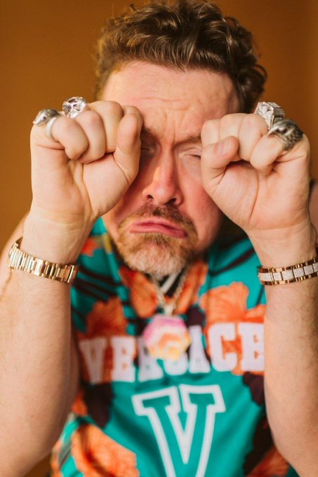 A man with a beard wearing a colorful floral Versace shirt and multiple gold rings and bracelets, rubbing his eyes with his fists.