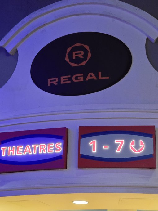 The image shows a movie theater with two signs above the entrance, one reading "Regal" and another displaying the showtimes for a movie called "Good Night". The sign for "Regal" is located at the top of the building, while the other sign is positioned below it. Both signs are illuminated in blue neon lights to attract attention from potential moviegoers.