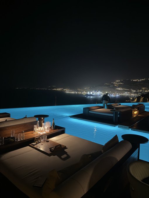 A luxurious infinity pool glows electric blue against the dark night sky, with elegant cabanas and low tables set up poolside. In the distance, the twinkling lights of a coastal town stretch along the shoreline, capturing the serene beauty of a nighttime retreat in Mykonos.