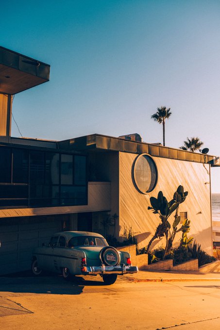 A vintage teal and white classic car parked on a sunlit street in front of a mid-century modern building with a distinctive round window and palm trees.