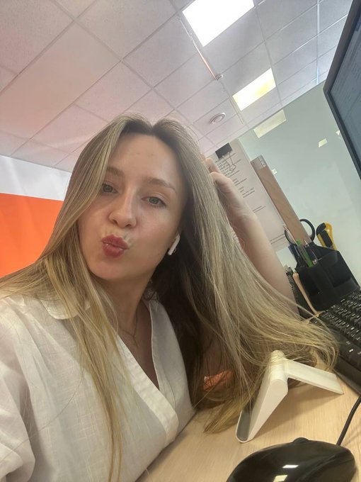 A blonde woman in a white blouse taking a selfie at an office desk while making a pouty face, wearing wireless earbuds with office equipment visible in the background.