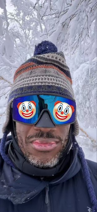 The image features a man wearing a hat with two clown faces on it, sunglasses, and goggles. The man appears to be outdoors, possibly in a snowy environment. There are also other people visible in the background of the photo.