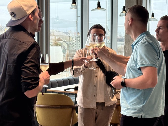 Four men raise glasses of wine in a toast at what appears to be a restaurant with city views, gathered together to celebrate Prague's longstanding role as a pioneer in the Bitcoin movement.