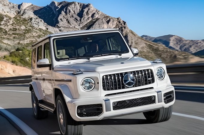 A white Mercedes-Benz G-Wagon SUV driving on a winding mountain road with rocky peaks in the background.