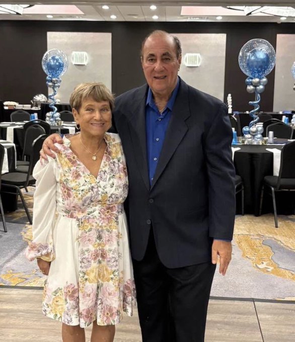 An older couple poses together at what appears to be a formal banquet hall decorated with blue and silver balloons.