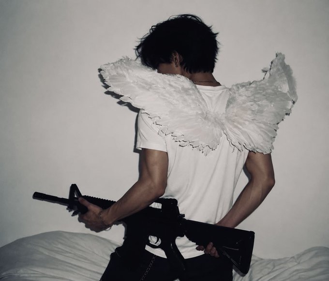 A person wearing white angel wings and a white t-shirt stands with their back to the camera, holding a black rifle.