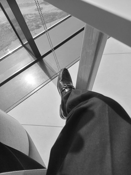 A black and white photo taken from a first-person perspective looking down at a person's foot in a leather loafer resting against a glass window frame on a high floor, showing the ground far below.
