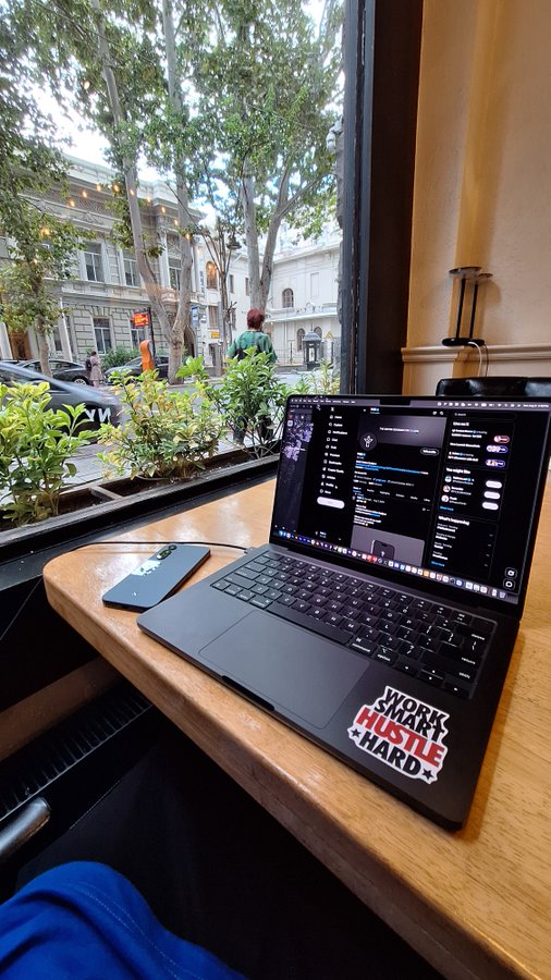 A MacBook displaying the Solana ecosystem interface sits on a wooden café table by a window, complete with a "WORK SMART HUSTLE HARD" sticker and a phone beside it, illustrating the post's theme of working and traveling within the crypto-native lifestyle.