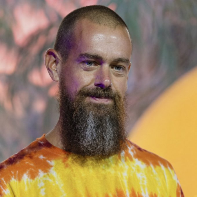 A close-up portrait of a bearded man with a shaved head wearing an orange and yellow tie-dye shirt, smiling at the camera.