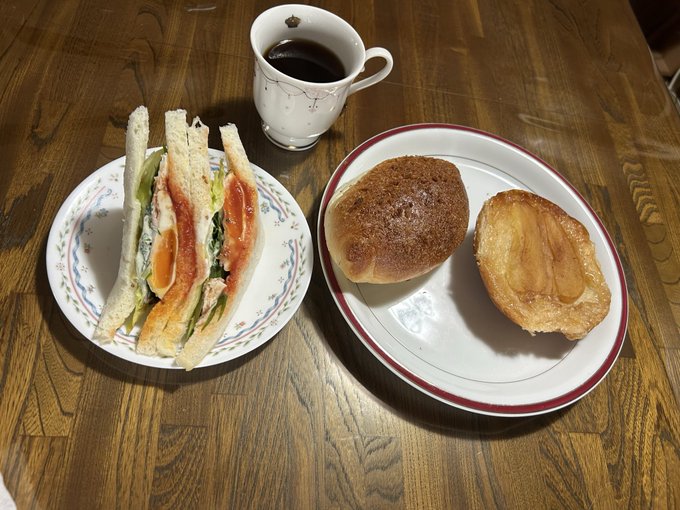 A simple meal on a wooden table consisting of a cut sandwich with egg and vegetables, a bread roll, a pastry, and a cup of black coffee.