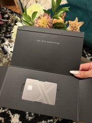 The image shows a gray X-branded credit card presented in a dark gray box with the text "TAP INTO EVERYTHING" visible inside the lid. This card, likely for the "everything app" X, is shown alongside a bouquet of flowers, hinting at the user's ability to use their XMoney yield to purchase everyday items like coffee.