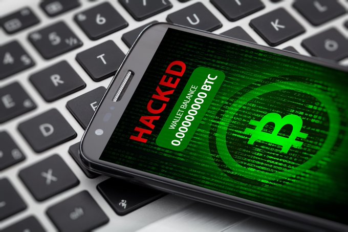 A smartphone resting on a keyboard displays a screen with the word 'HACKED' in red alongside a Bitcoin symbol and a wallet balance of 0.00000000 BTC, set against a green digital code background.