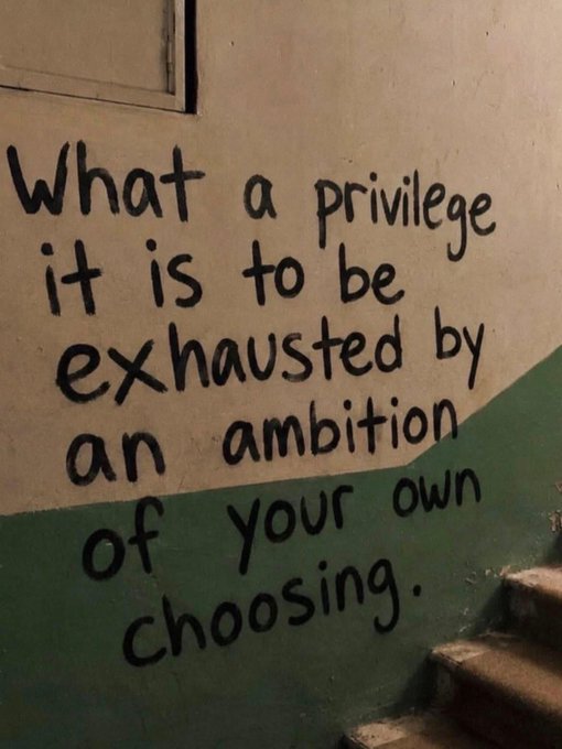 Hand-painted graffiti on a wall that reads: 'What a privilege it is to be exhausted by an ambition of your own choosing.'